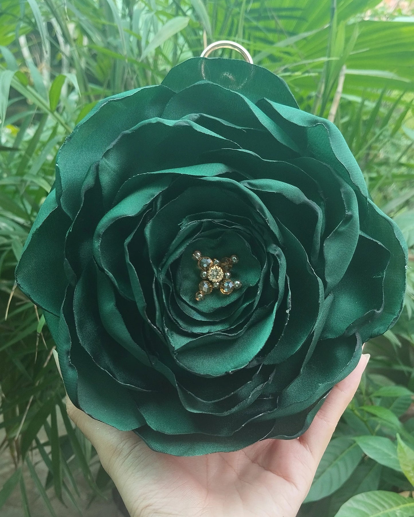 A hand holding a large, dark green fabric rose ornament with a small gold star detail in the center, set against a background of green foliage.