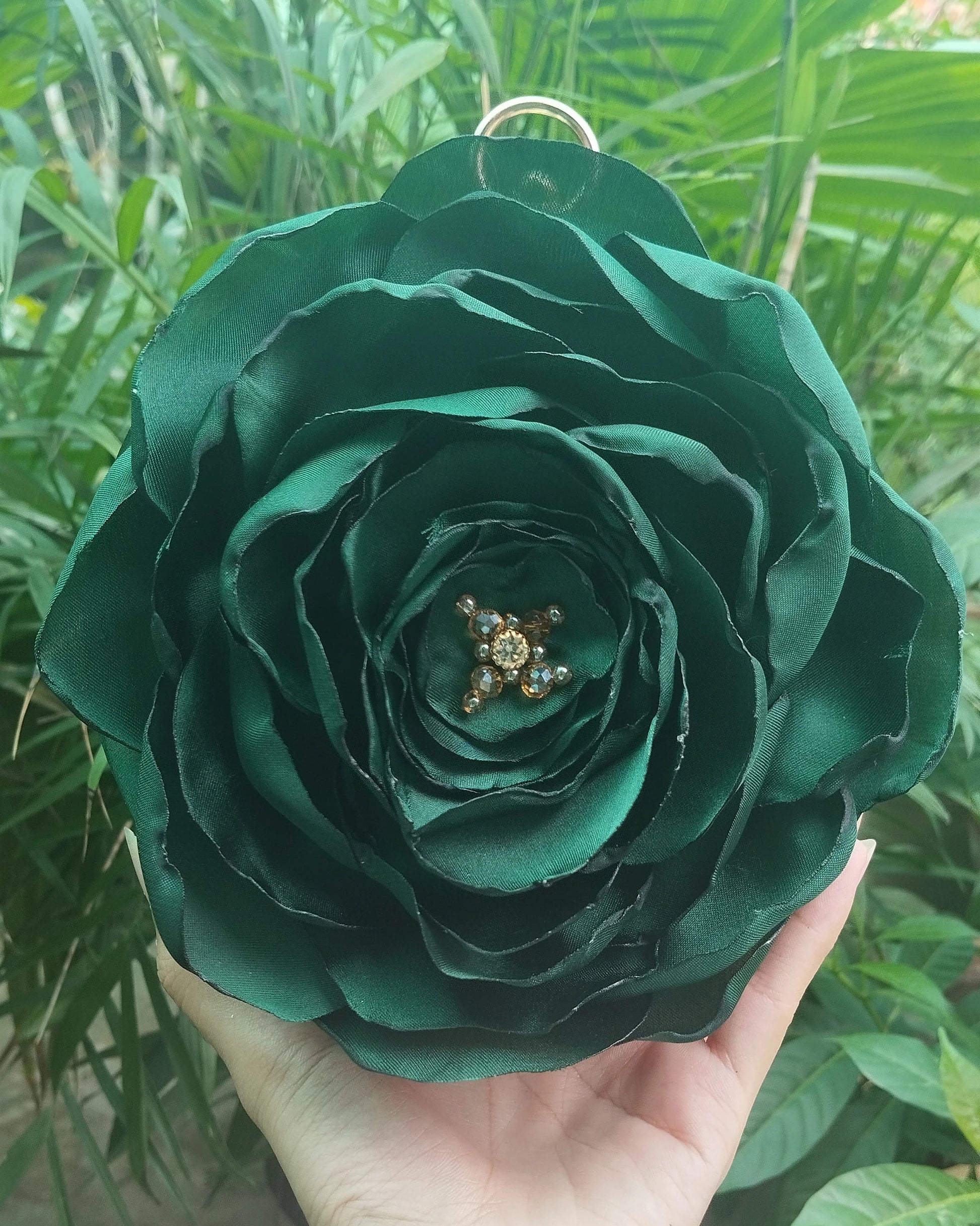 A hand holding a large, dark green fabric rose ornament with a small gold star detail in the center, set against a background of green foliage.