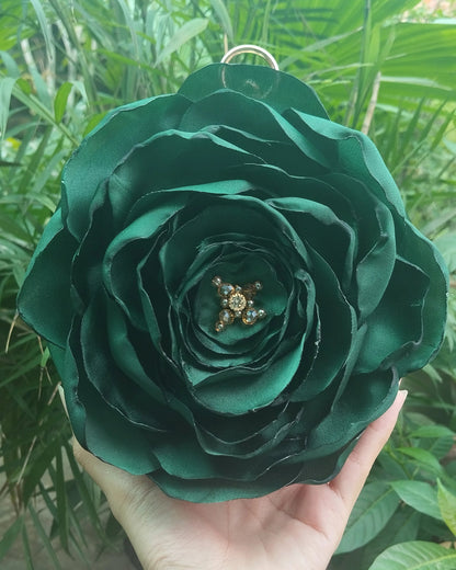 A hand holding a large, dark green fabric rose ornament with a small gold star detail in the center, set against a background of green foliage.