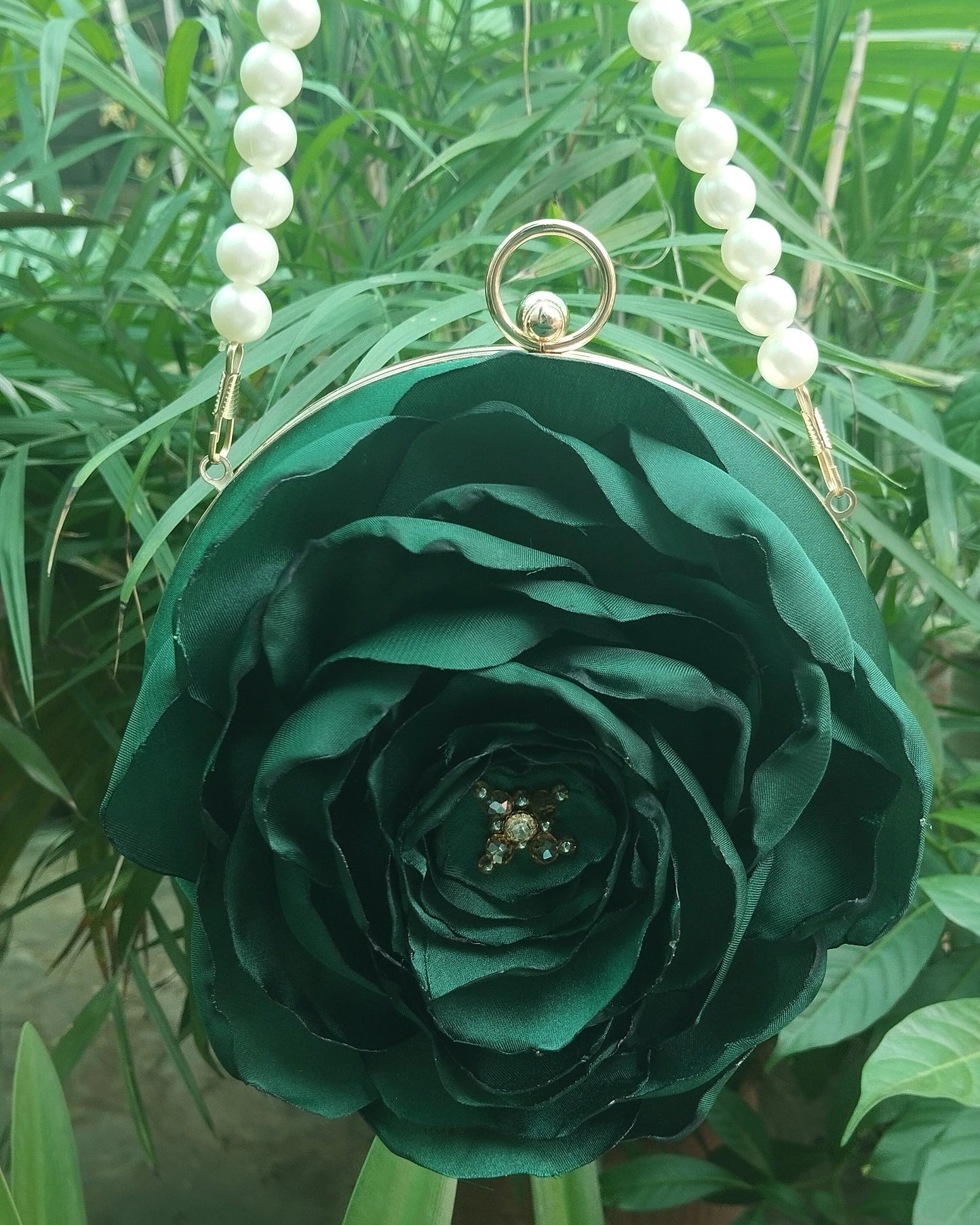 A dark green fabric rose-shaped purse with pearl bead shoulder straps and a gold-tone ring clasp, photographed outdoors against a background of green foliage.