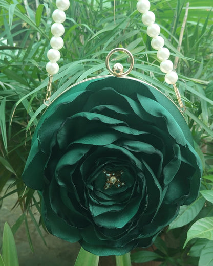 A dark green fabric rose-shaped purse with pearl bead shoulder straps and a gold-tone ring clasp, photographed outdoors against a background of green foliage.