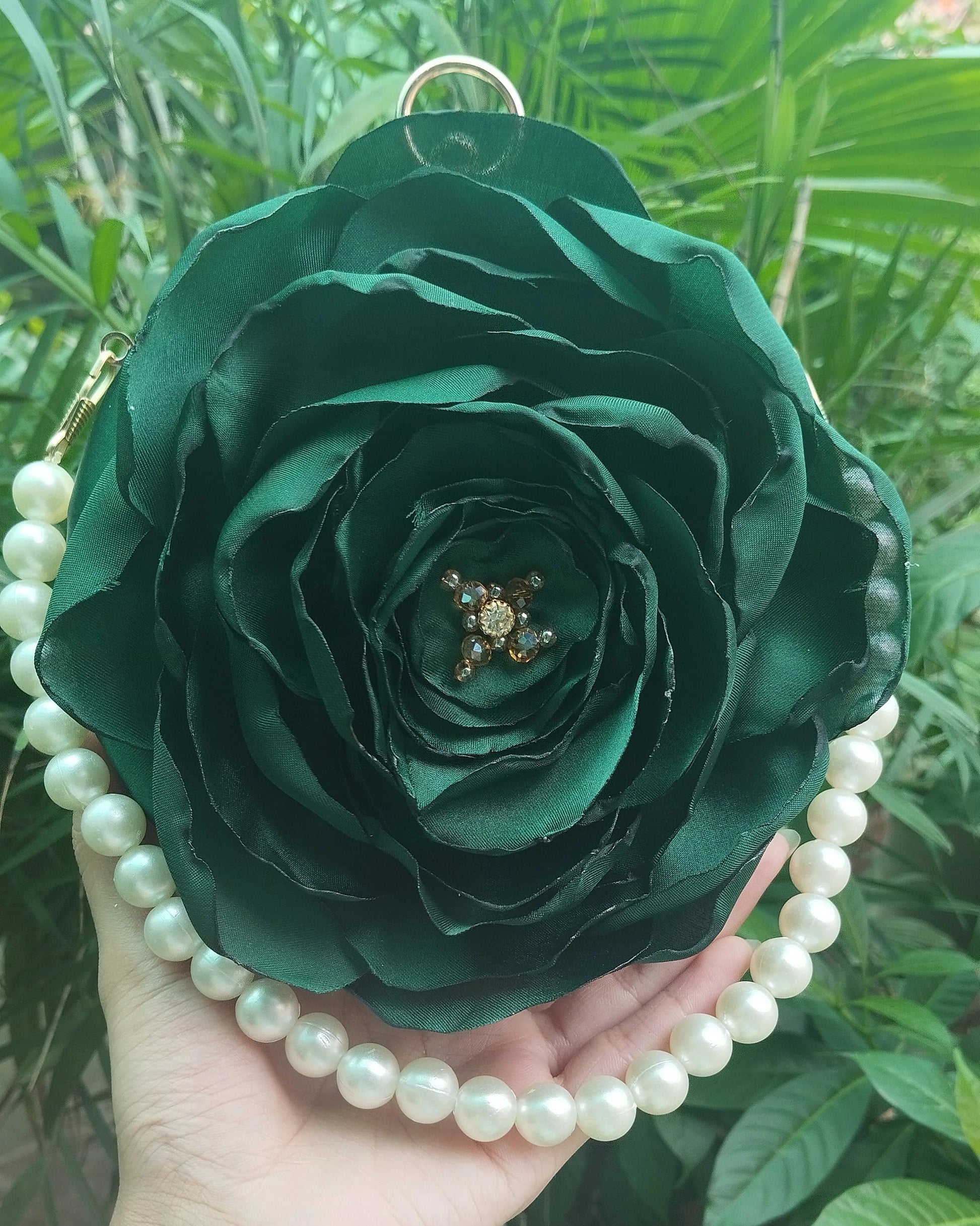 A dark green satin rose-shaped purse with a pearl bead border and a small gold-toned brooch in the center, held in a hand against a background of green foliage.