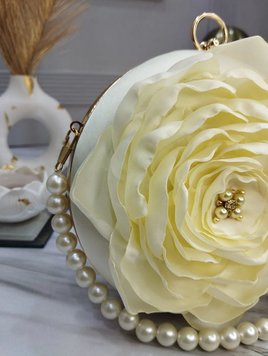 A cream-colored satin flower purse with a gold-tone metal frame, adorned with a pearl bead strap and a central gold-tone floral embellishment.